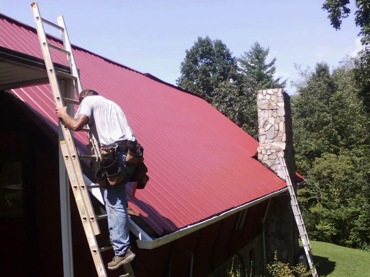 Roofer on a ladder beside a red metal roof and stone chimney — Soffit & Fascia Repair by Roofing Brothers in Deary